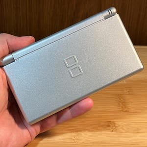 May include: A silver handheld gaming console with a rectangular, clamshell design. The top cover features a metallic finish and a centered, embossed DS logo. The device has a hinge along the top edge and a smooth, clean surface texture throughout.