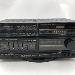 Puede incluir: Receptor estéreo vintage negro con múltiples paneles de control, incluyendo un sintonizador, un ecualizador gráfico y un temporizador digital. El receptor tiene varios botones, perillas y una pantalla. La marca "Sansui" es visible.