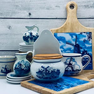 Può includere: Una collezione di ceramiche blu e bianche di Delft, tra cui una saliera, piccole brocche, una tazza e un barattolo. I pezzi presentano disegni di mulini a vento e paesaggi. È presente anche un tagliere in legno con una piastrella di Delft.