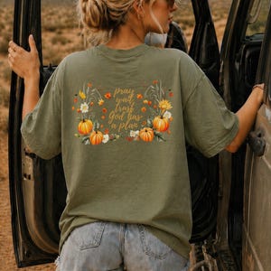 May include: An olive green short-sleeve t-shirt featuring a back graphic of orange pumpkins, yellow sunflowers, and autumn leaves. The text in the center reads, "pray wait trust God has a plan" in a cursive brown font.