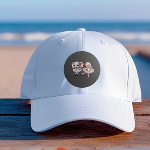 Puede incluir: Gorra de béisbol blanca con un parche redondo gris oscuro. El parche presenta un elefante y un pollito con atuendos de ballet, con el texto "DANCE FOR JOY". La gorra está hecha de un material liso.