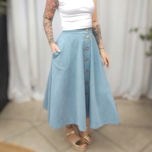 Könnte beinhalten: Hellblauer Jeansrock mit großen Holzknöpfen auf der Vorderseite. Der Rock hat eine ausgestellte A-Linien-Form und Taschen. Das Model trägt ein weißes Oberteil mit kurzen Ärmeln und goldene Sandalen.
