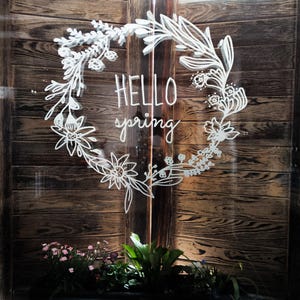 Könnte beinhalten: Ein weißer Blumenkranz, der auf eine Holztür gezeichnet ist, mit dem Text "HELLO spring" im Kranz.