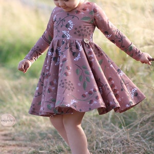 May include: A young girl wearing a long-sleeved, pink dress with a floral print. The dress has a gathered skirt and a button closure at the neckline. The girl is standing in a field of tall grass.