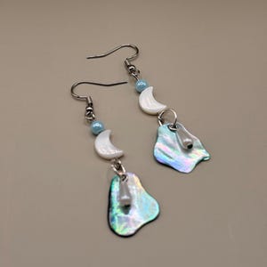 May include: Dangle earrings with iridescent, teardrop-shaped pendants. Each earring has a small, white crescent moon and a light blue bead. The earrings are suspended from silver-coloured ear wires.