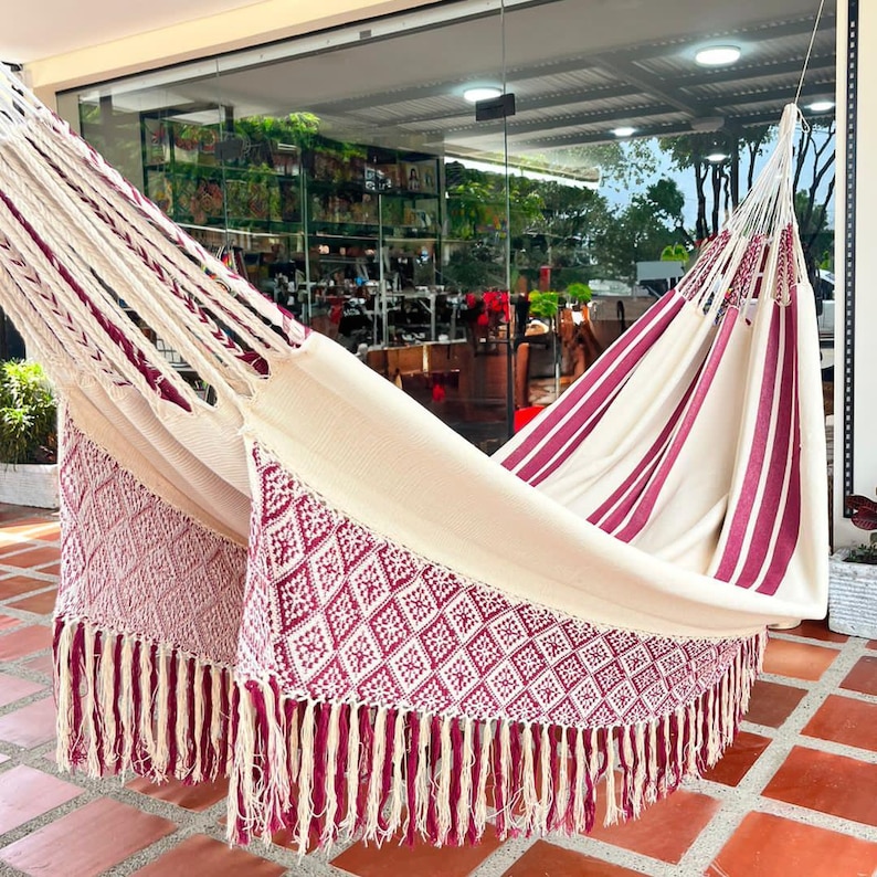 Puede incluir: Una hamaca color crema con detalles burdeos. La hamaca presenta un dise&ntilde;o tejido con patrones de diamantes y flecos. Rayas burdeos adornan la tela de la hamaca, creando un contraste visualmente atractivo. La hamaca est&aacute; dise&ntilde;ada para la relajaci&oacute;n y el uso al aire libre.