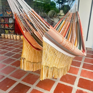 Puede incluir: Dos coloridas hamacas con patrones tejidos y bordes con flecos cuelgan al aire libre. Una hamaca presenta rayas naranjas, blancas y marrones, mientras que la otra tiene patrones amarillos y blancos. Las hamacas est&aacute;n suspendidas de cuerdas blancas.
