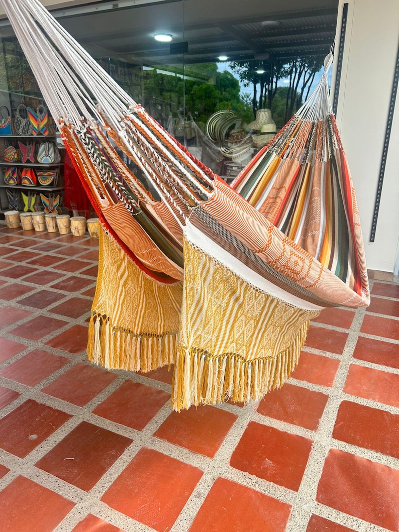 Puede incluir: Dos coloridas hamacas con patrones tejidos y bordes con flecos cuelgan al aire libre. Una hamaca presenta rayas naranjas, blancas y marrones, mientras que la otra tiene patrones amarillos y blancos. Las hamacas est&aacute;n suspendidas de cuerdas blancas.
