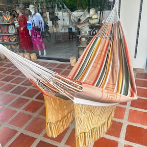 Puede incluir: Una colorida hamaca con dise&ntilde;o a rayas en tonos naranja, verde y blanco. La hamaca tiene un borde con flecos en amarillo y blanco. La hamaca est&aacute; colgada frente a una tienda.