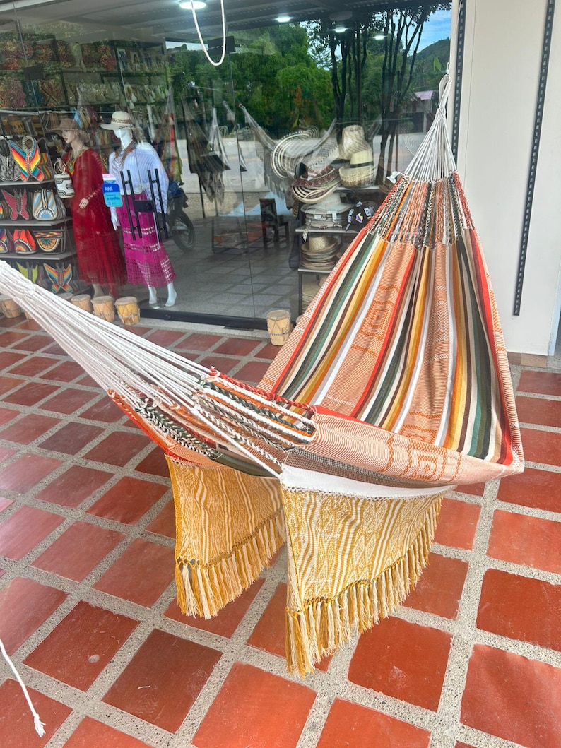 Puede incluir: Una colorida hamaca con dise&ntilde;o a rayas en tonos naranja, verde y blanco. La hamaca tiene un borde con flecos en amarillo y blanco. La hamaca est&aacute; colgada frente a una tienda.