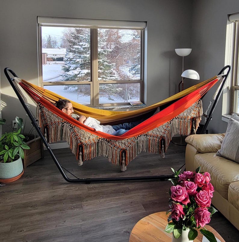 Puede incluir: Una colorida hamaca sobre un soporte de metal negro. La hamaca presenta un dise&ntilde;o vibrante con tonos de amarillo, naranja y rojo, y est&aacute; adornada con flecos decorativos. La escena se desarrolla en interiores con una vista nevada al aire libre visible a trav&eacute;s de una gran ventana.