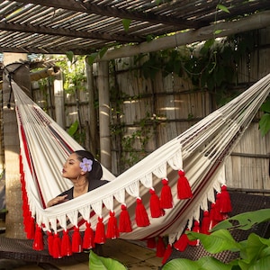 Puede incluir: Una hamaca crema con detalles y borlas rojas cuelga al aire libre. Una persona se relaja dentro. La hamaca est&aacute; suspendida entre soportes, con una p&eacute;rgola de madera y exuberante vegetaci&oacute;n como tel&oacute;n de fondo.
