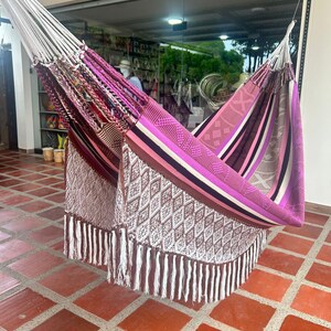 Puede incluir: Una colorida hamaca con un dise&ntilde;o tejido, con tonos de rosa, morado, marr&oacute;n y blanco. La hamaca tiene un borde con flecos y est&aacute; suspendida de cuerdas blancas. El dise&ntilde;o incluye patrones geom&eacute;tricos y rayas.