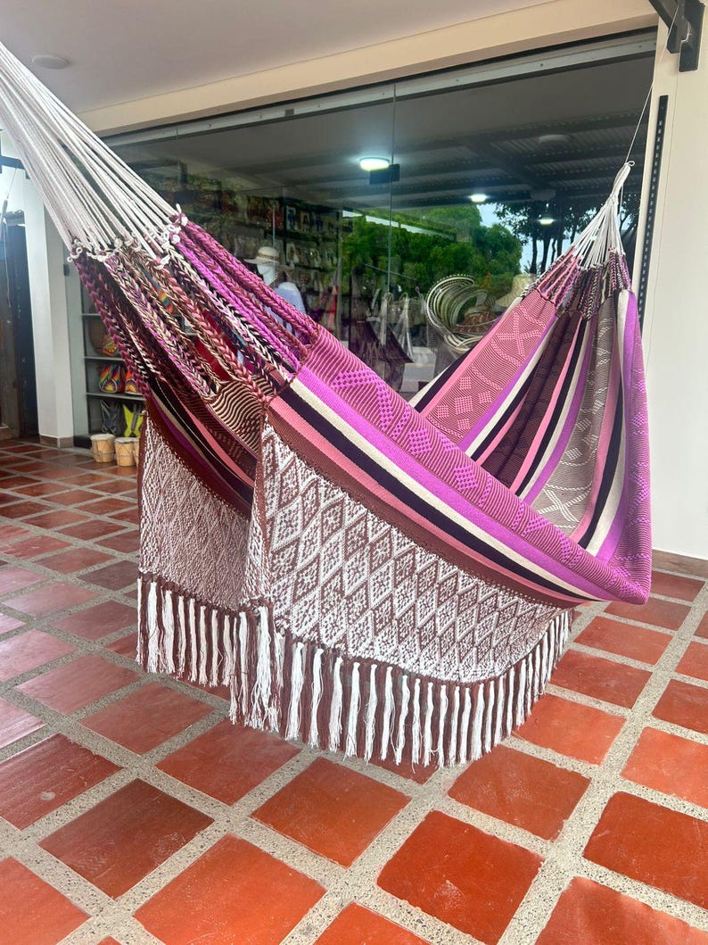 Puede incluir: Una colorida hamaca con un dise&ntilde;o tejido, con tonos de rosa, morado, marr&oacute;n y blanco. La hamaca tiene un borde con flecos y est&aacute; suspendida de cuerdas blancas. El dise&ntilde;o incluye patrones geom&eacute;tricos y rayas.