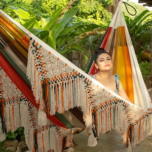 Puede incluir: Una colorida hamaca con detalles de macram&eacute; y flecos. La hamaca presenta una mezcla de rayas naranjas, rojas, verdes y amarillas. Est&aacute; colgada al aire libre, con un exuberante follaje verde de fondo.