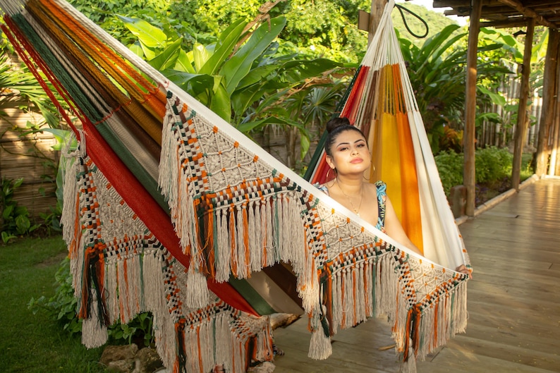 Puede incluir: Una colorida hamaca con detalles de macram&eacute; y flecos. La hamaca presenta una mezcla de rayas naranjas, rojas, verdes y amarillas. Est&aacute; colgada al aire libre, con un exuberante follaje verde de fondo.