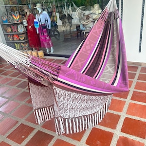 Puede incluir: Una colorida hamaca con un dise&ntilde;o a rayas en tonos de rosa, morado, marr&oacute;n y blanco. La hamaca tiene un patr&oacute;n tejido con flecos. La hamaca se exhibe al aire libre sobre una superficie embaldosada.