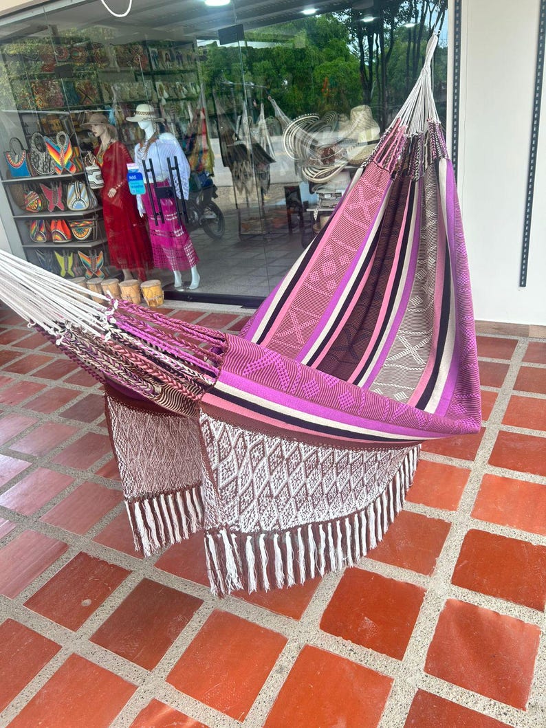 Puede incluir: Una colorida hamaca con un dise&ntilde;o a rayas en tonos de rosa, morado, marr&oacute;n y blanco. La hamaca tiene un patr&oacute;n tejido con flecos. La hamaca se exhibe al aire libre sobre una superficie embaldosada.