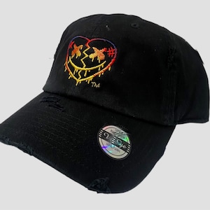 May include: Black distressed baseball cap with a colourful embroidered heart design. The heart is cracked and dripping with yellow and orange paint. The text "TM" is embroidered below the heart.