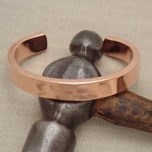 Op de afbeelding: Een koperen manchetarmband met een gehamerde textuur, rustend op een metalen hamer. De armband heeft een eenvoudig, open ontwerp en een gepolijste afwerking. De hamer heeft een houten handvat.