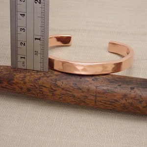 May include: A hammered copper bracelet measuring 2.5 cm in diameter. The bracelet is shown on a wooden surface.
