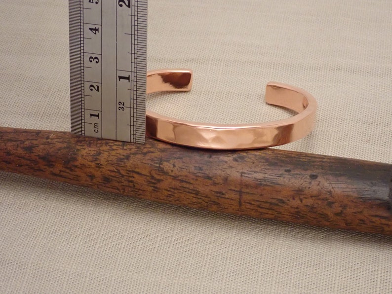 May include: A hammered copper bracelet measuring 2.5 cm in diameter. The bracelet is shown on a wooden surface.