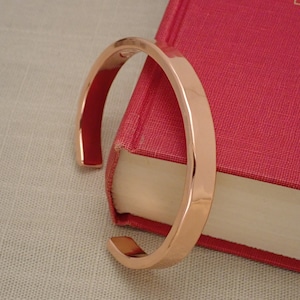 May include: A rose gold bangle bracelet with a smooth, polished finish. The bracelet is open and rests on a red book.