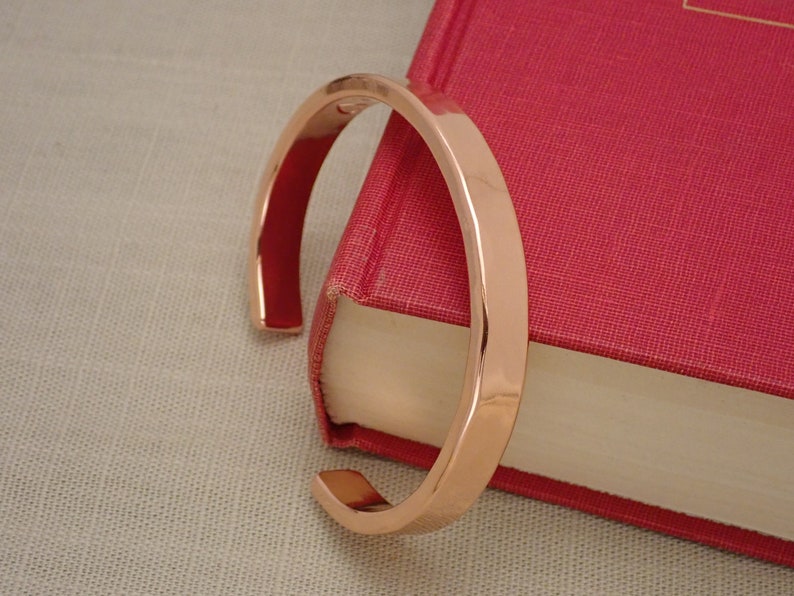 May include: A rose gold bangle bracelet with a smooth, polished finish. The bracelet is open and rests on a red book.