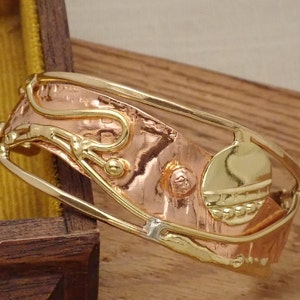 May include: A gold and copper bracelet with a textured design and a circular accent. The bracelet is made of metal and has a unique, handcrafted look.