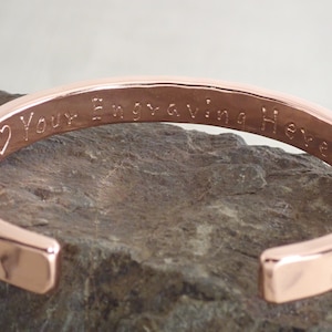 May include: A rose gold bangle bracelet with a heart and the text "Your Engraving Here" engraved on the inside.