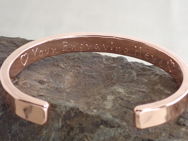 May include: A rose gold bangle bracelet with a heart and the text "Your Engraving Here" engraved on the inside.