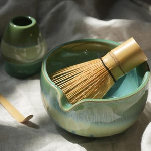 May include: A matcha tea set featuring a green ceramic bowl with a bamboo whisk, a small green ceramic holder, and a bamboo scoop. The bowl has a turquoise interior and a light green exterior. The logo "Waisked." is in the upper right corner.