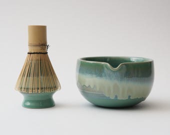 May include: A matcha tea set featuring a green ceramic bowl with a spout and a bamboo whisk. The bowl has a gradient of green and white, while the whisk is made of thin bamboo bristles.