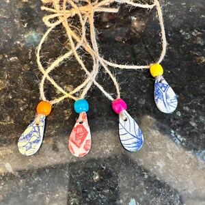 May include: Four handmade teardrop-shaped pendants with colorful beads and twine. Each pendant has a unique pattern in red, blue, and yellow. The pendants are strung on natural twine, creating a rustic and artistic look.