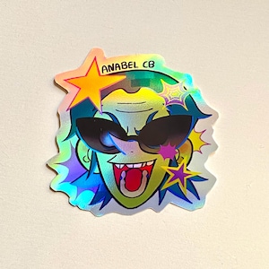 May include: Holographic sticker featuring a cartoon face with sunglasses, a wide open mouth, and spiky hair. The sticker has a star and the text "ANABEL CB". The sticker has a rainbow effect.