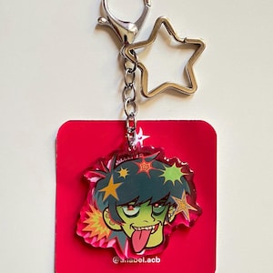May include: A red acrylic keychain featuring a cartoon character with green skin, spiky green hair, and a tongue sticking out. The keychain has a silver star-shaped charm and a lobster clasp. The text "@ariabel.acb" is visible.