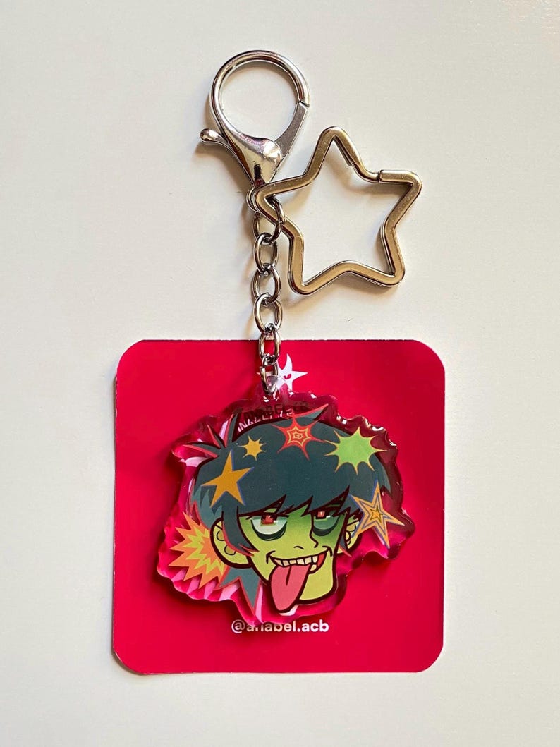 May include: A red acrylic keychain featuring a cartoon character with green skin, spiky green hair, and a tongue sticking out. The keychain has a silver star-shaped charm and a lobster clasp. The text "@ariabel.acb" is visible.