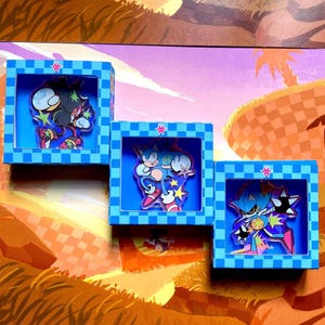 Puede incluir: Tres cajas cuadradas de cuadros azules, cada una con una figura de personaje de acrílico. La caja de la izquierda contiene un personaje oscuro, la central un personaje azul y la de la derecha un personaje azul metálico con púas.