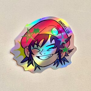 May include: Holographic sticker featuring a smiling cartoon character with purple hair and a pink hat. The sticker has green and purple star accents and the text "ANABEL CB". The sticker has a shiny, iridescent finish.