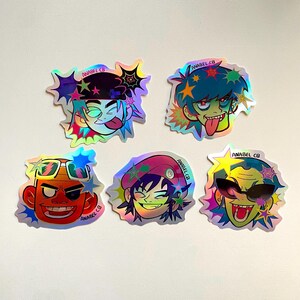 May include: A collection of five holographic stickers featuring cartoon character designs. Each sticker has a unique character with vibrant colors, including red, blue, green, and yellow. The stickers have a starburst design and the text "ANABEL CB".