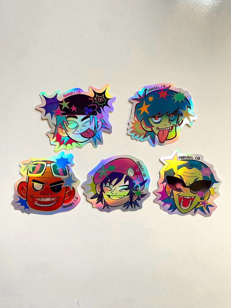 May include: A collection of five holographic stickers featuring cartoon character designs. Each sticker has a unique character with vibrant colors, including red, blue, green, and yellow. The stickers have a starburst design and the text "ANABEL CB".