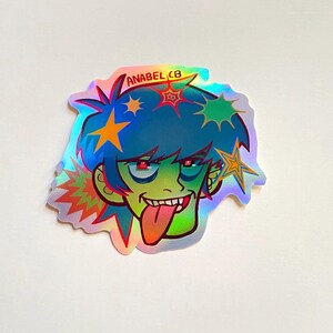 May include: Holographic sticker featuring a cartoon character with blue hair, green skin, and a red tongue sticking out. The character has red eyes and is decorated with star-shaped accents. The sticker has the text "ANABEL CB".