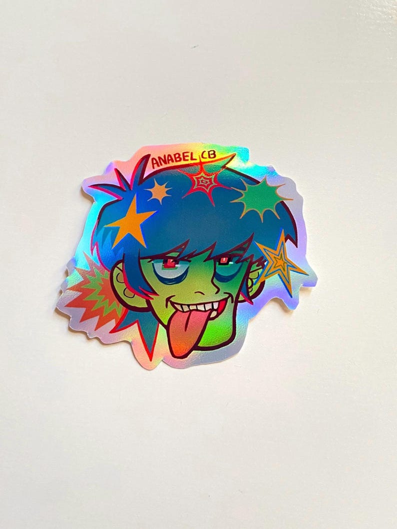 May include: Holographic sticker featuring a cartoon character with blue hair, green skin, and a red tongue sticking out. The character has red eyes and is decorated with star-shaped accents. The sticker has the text "ANABEL CB".