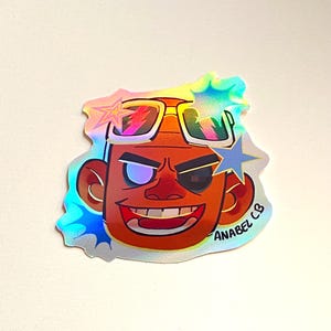 May include: A holographic sticker featuring a cartoon face with sunglasses and a star-shaped design. The sticker has the text "ANABEL CB" and is in shades of orange, red, and blue.
