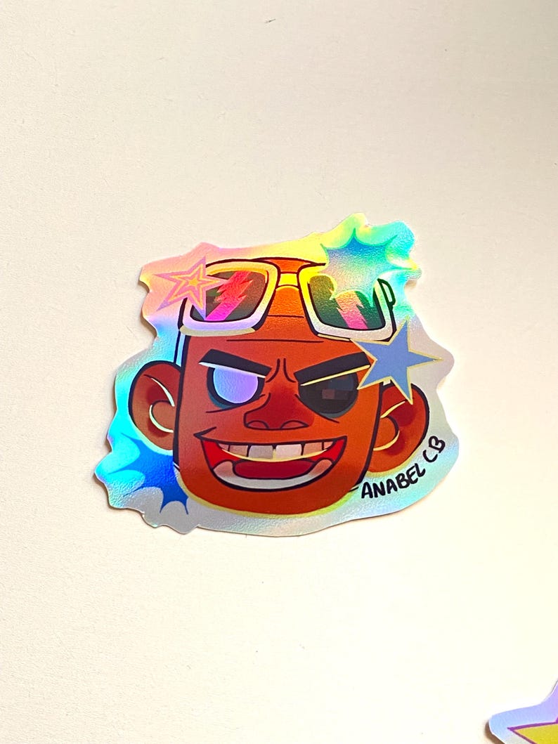 May include: A holographic sticker featuring a cartoon face with sunglasses and a star-shaped design. The sticker has the text "ANABEL CB" and is in shades of orange, red, and blue.