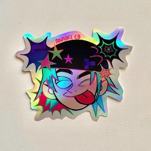 May include: Holographic sticker featuring a cartoon character with blue hair, a black hat, and a playful expression. The sticker has a unique shape with star and web-like accents. The text "ANABEL CB" is visible.