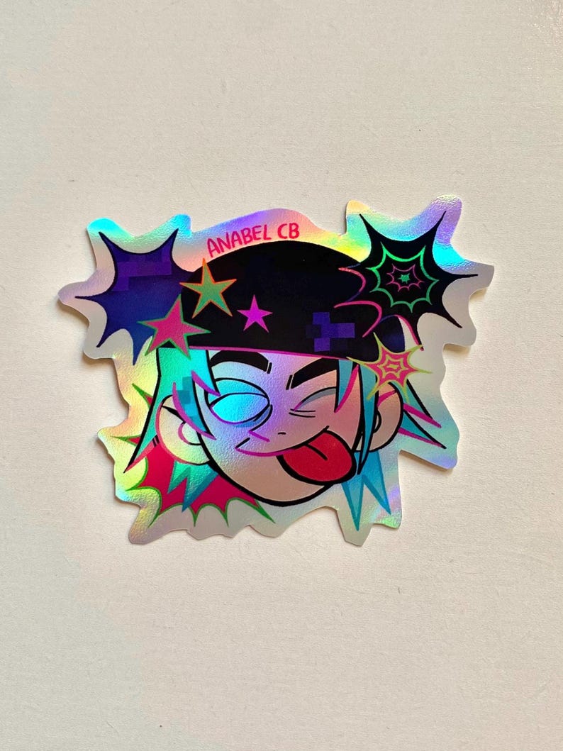 May include: Holographic sticker featuring a cartoon character with blue hair, a black hat, and a playful expression. The sticker has a unique shape with star and web-like accents. The text "ANABEL CB" is visible.