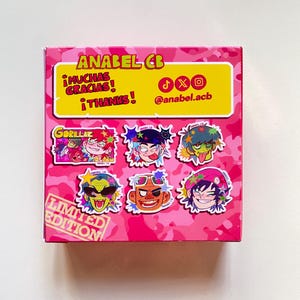 May include: A pink box with a camouflage pattern featuring cartoon character stickers. The box has text that reads "ANABEL CB", "&iexcl;MUCHAS GRACIAS! &iexcl;THANKS!", and "LIMITED EDITION". The stickers feature colorful illustrations of the Gorillaz band members.