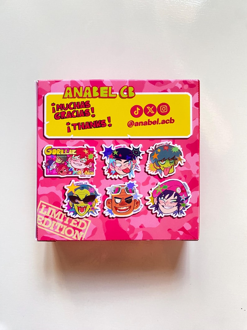 May include: A pink box with a camouflage pattern featuring cartoon character stickers. The box has text that reads "ANABEL CB", "&iexcl;MUCHAS GRACIAS! &iexcl;THANKS!", and "LIMITED EDITION". The stickers feature colorful illustrations of the Gorillaz band members.
