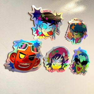 May include: A collection of six holographic stickers featuring cartoon character faces with various expressions. The stickers have a starburst design around the edges and the text "ANABEL CB" is printed on each sticker.